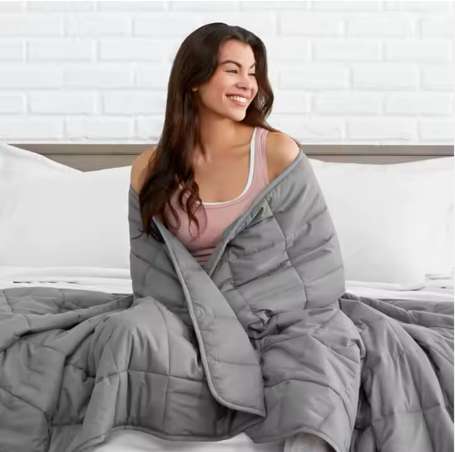 A smiling woman wrapped in a cozy gray weighted blanket, sitting on a white bed with white pillows and a white brick wall background. The blanket appears soft and quilted, providing comfort and warmth.