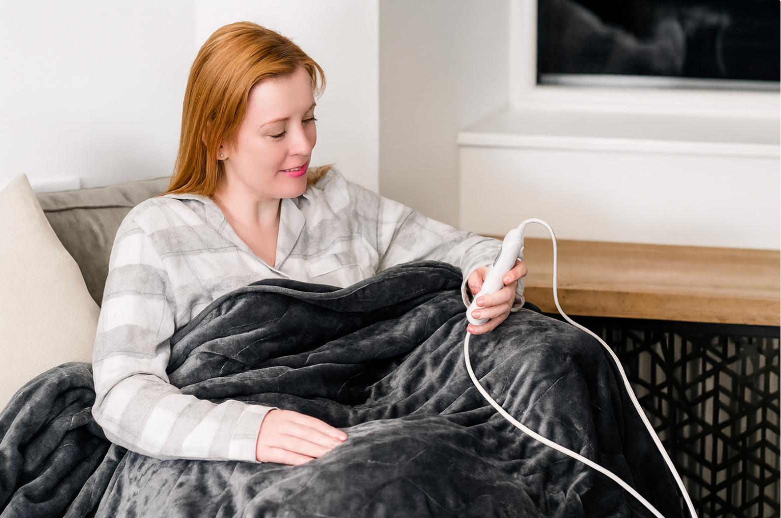 Woman relaxing on a couch with a soft grey electric blanket, adjusting the heat settings using the controller
