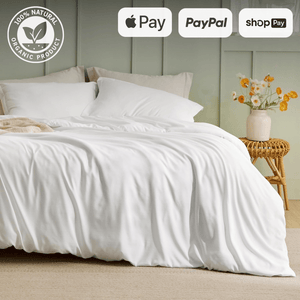 Luxurious white bed with soft, draped sheets in a cozy bedroom setting. The bed is adorned with pillows and a light beige throw blanket. A vase with fresh flowers sits on a wicker side table, adding a touch of nature to the room. The image includes logos for Apple Pay, PayPal, and Shop Pay, highlighting convenient payment options. The '100% Natural Organic Product' Badgee emphasizes the eco-friendly aspect of the bedding