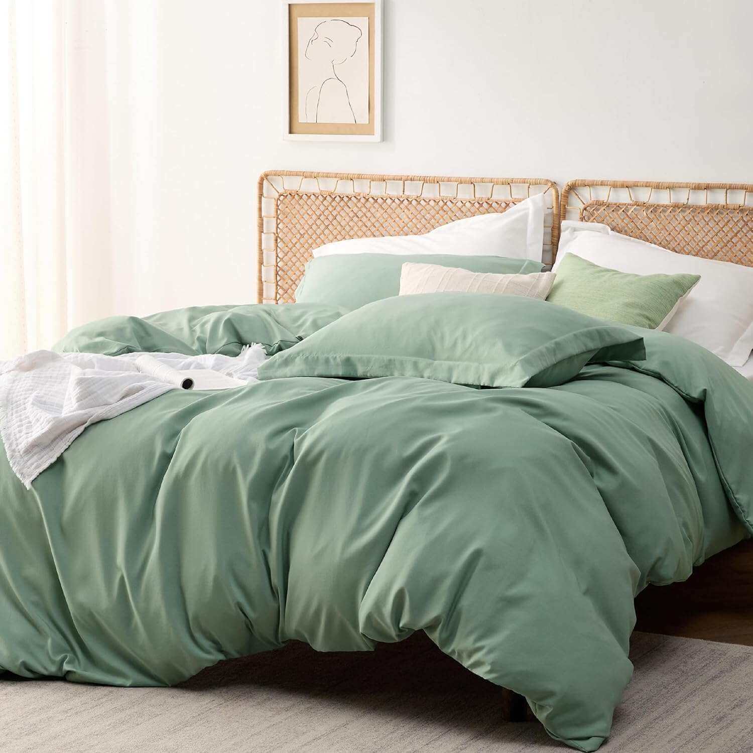 Sage green bedding set on a bed with white sheets and a textured white throw, near a window with sheer curtains and rattan headboard